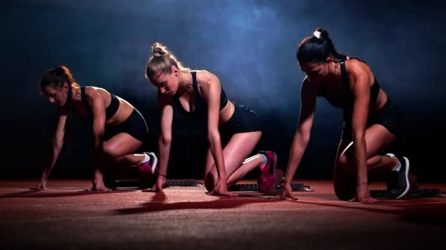 Sport:-drei-Mädchen-in-schwarzen-Klamotten-des-Sportlers-in-der-Nacht-auf-dem-Laufband-startet-ein-Rennen-auf-der-Sprintdistanz-aus-der-Sitzposition