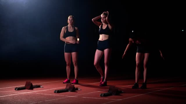 Sport:-drei-Mädchen-in-schwarzen-Klamotten-des-Sportlers-in-der-Nacht-auf-dem-Laufband-startet-ein-Rennen-auf-der-Sprintdistanz-aus-der-Sitzposition