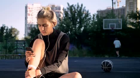 Gorgeous-blonde-woman-listening-music-through-earphones-and-typing-on-phone,-sitting-on-court,-man-in-background-throwing-ball