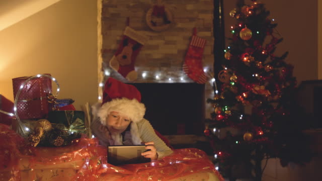 Glückliches-kleines-Mädchen-mit-Tablet-auf-einen-Videoanruf-zu-Weihnachten-in-eingerichteten-Wohnzimmer-mit-Baum-und-Schornstein.