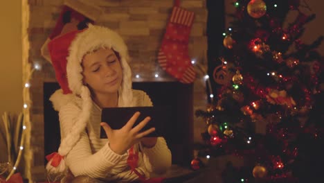 Glückliches-kleines-Mädchen-mit-Tablet-auf-einen-Videoanruf-zu-Weihnachten-in-eingerichteten-Wohnzimmer-mit-Baum-und-Schornstein.