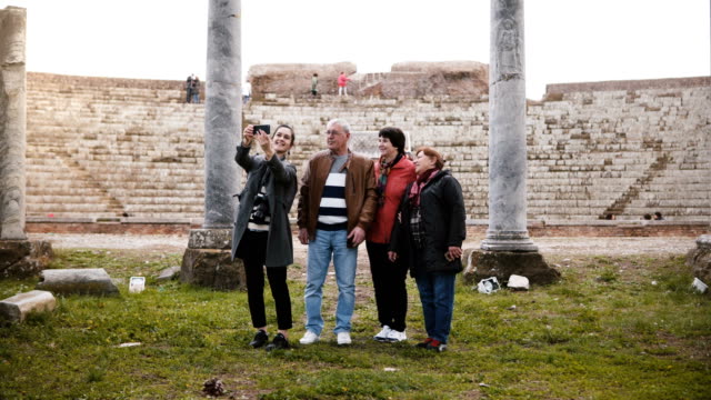 Hombre-senior-feliz-y-las-mujeres-con-niña-agitando-en-video-chat-con-familiares-cerca-de-antiguas-ruinas-del-anfiteatro-en-Ostia,-Italia.