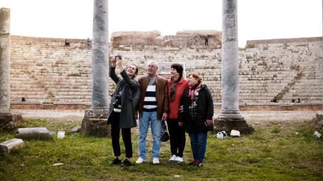 Lächelnde-junge-europäische-Mädchen-und-glücklich-senior-Reisegruppe-unter-Selfie-in-der-Nähe-von-alten-Amphitheater-Ruinen-in-Ostia,-Italien.