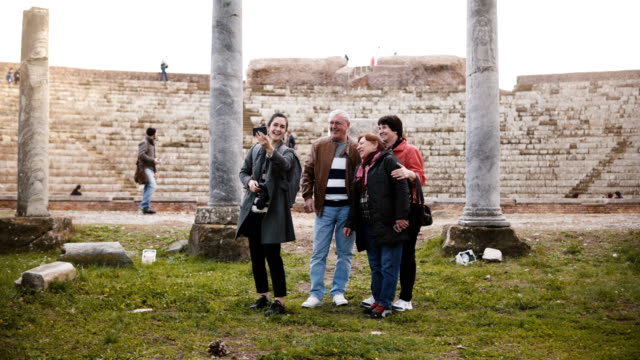 Smiling-senior-family-and-young-woman-tourists-waving-on-video-call-to-family-at-old-amphitheater-ruins-in-Ostia,-Italy.