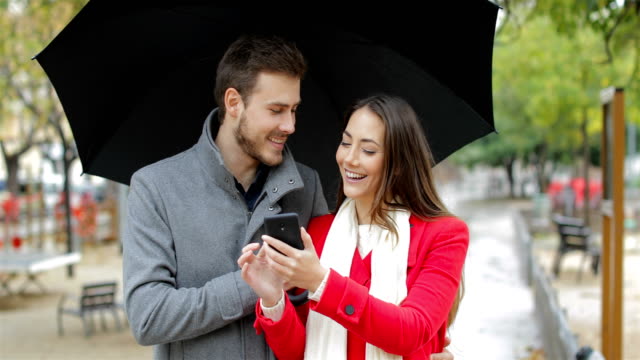 Happy-couple-checking-smart-phone-under-the-rain