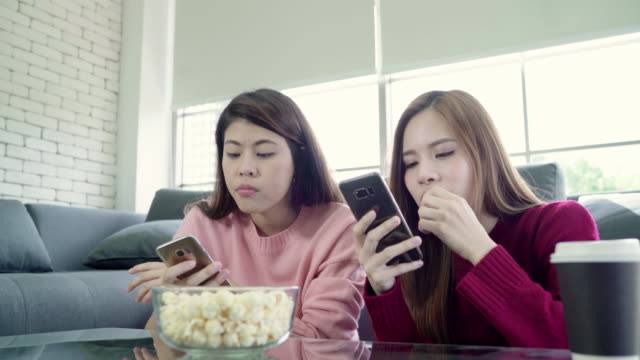 Las-mujeres-asiáticas-con-smartphone-y-comer-palomitas-de-maíz-en-la-sala-de-estar-en-casa,-grupo-de-amigo-de-compañero-de-cuarto-disfrutan-momento-divertido-mientras-se-está-acostado-en-el-sofá.-Las-mujeres-de-estilo-de-vida-en-casa-relajación-conce