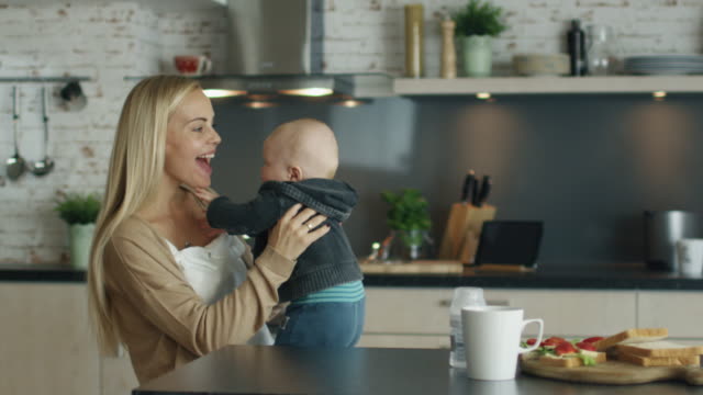 Young-Mother-Plays-with-Her-Beautiful-Baby-while-Standing-on-the-Kitchen.-In-Slow-Motion.