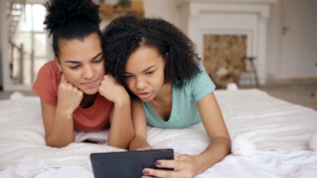 Zwei-fröhliche-gemischte-Rasse-lustige-Freundinnen-teilen-Soziale-Medien-mit-Tablet-Computer-und-Gespräche-liegen-im-Bett-zu-Hause