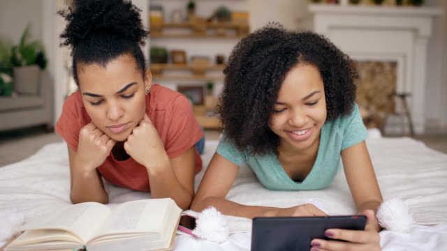 Two-cheerful-mixed-race-funny-girlfriends-reading-book-and-using-tablet-computer-talks-and-have-fun-lying-in-bed-at-home