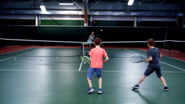 Ein-weibliche-Trainer-im-Sport-Anzug-lehrt-Tennisspielen-in-zwei-mittleren-Alters-Jungs,-Jungs-sind-Kugeln-auf-einem-Tennisplatz-Prellen