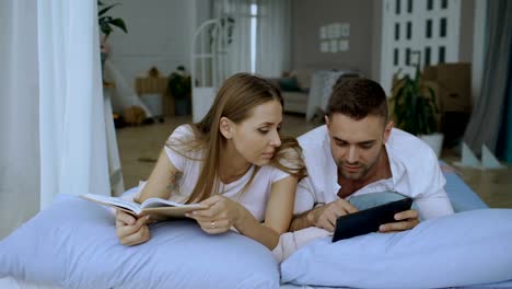Hübscher-junger-Mann-mit-Tablet-Computer-zeigen-Fotos-auf-dem-Tablet-PC-auf-seine-Girfriend-Buch-lesen-und-liegend-im-Bett-zu-Hause