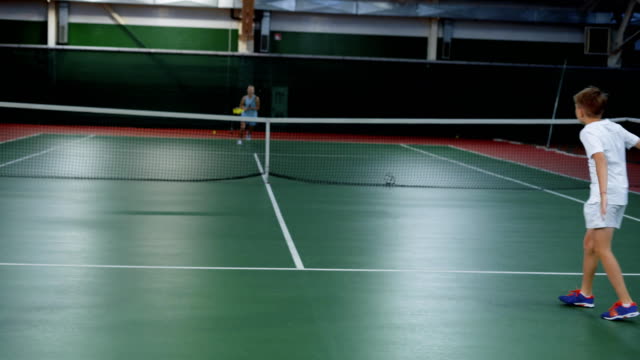 Two-young-athletes-jumping-in-recreation-area-playing-sport-game.-Three-happy-children-having-tennis-lesson-improving-skills.-Two-girls-and-boy-serving-and-returning-yellow-balls-with-rackets