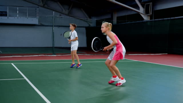 Two-young-athletes-in-recreation-area-playing-sport-game.-Happy-sister-and-brother-having-tennis-lesson-spending-time-at-indoor-court.-Girl-and-boy-serving-and-returning-yellow-balls-with-rackets