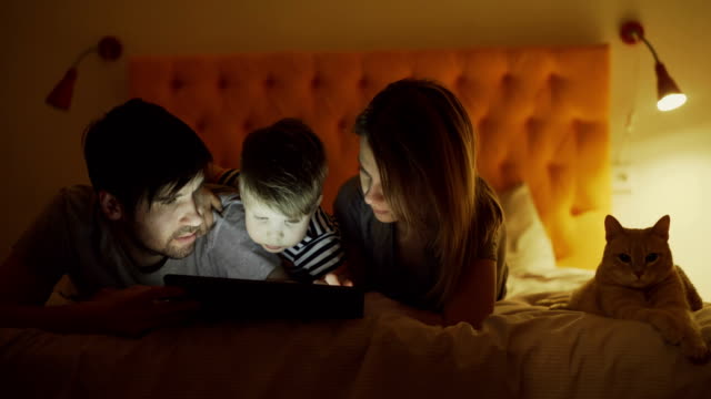 Glückliche-Familie-mit-kleinen-Sohn-und-lustige-Katze-liegend-im-Bett-zu-Hause-und-Surfen-social-Media-auf-dem-Tablet-PC-vor-dem-schlafen