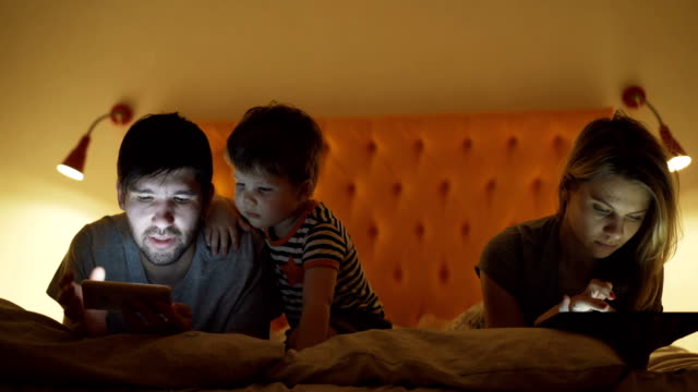 Glückliche-Familie-mit-kleinen-Sohn-im-Bett-zu-Hause-mittels-Tablet-PC-und-Smartphone-für-beobachtete-Film-und-Comic-abends-vor-dem-schlafen