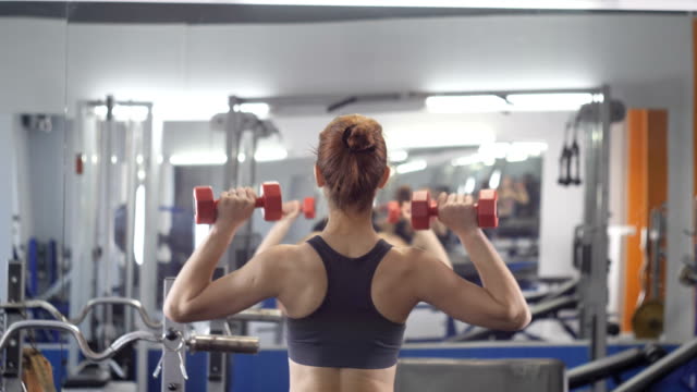 Young-beautiful-sporty-girl-doing-shoulder-press-with-dumbbells-exercising-in-a-sport-gym,-back-side-view