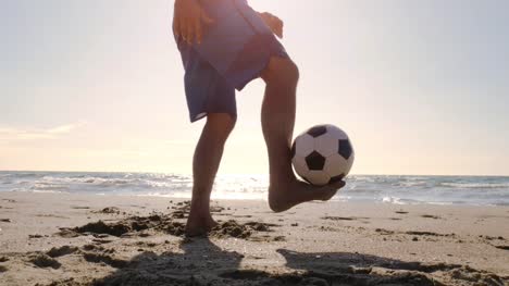 Hombre-joven-en-bañador-haciendo-trucos-con-fútbol-en-el-pie-en-la-orilla-del-mar-en-la-playa-de-silueta-al-atardecer