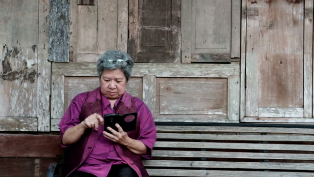 asian-elder-woman-holding-mobile-phone-beside-old-wood-house.-elderly-female-texting-message,-using-app-with-smartphone-in-park.-senior-use-cellphone-to-connect-with-social-network
