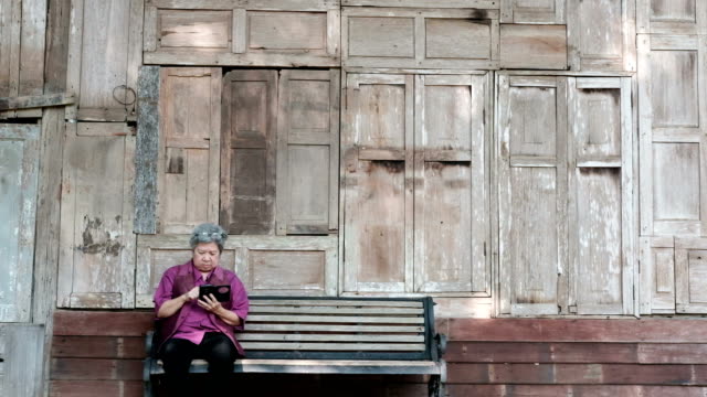 asian-elder-woman-holding-mobile-phone-beside-old-wood-house.-elderly-female-texting-message,-using-app-with-smartphone-in-park.-senior-use-cellphone-to-connect-with-social-network