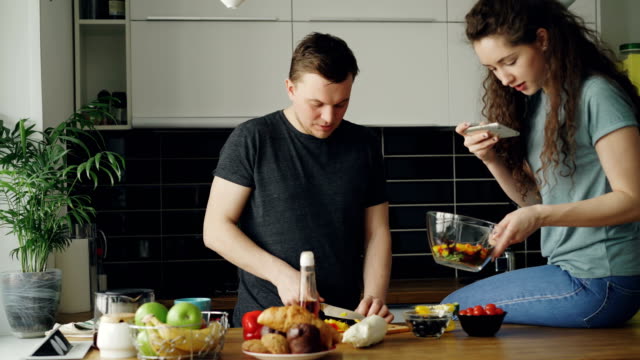 Attraktives-junges-Paar-Kochen-und-plaudern-gerne-in-der-Küche-zu-Hause.-Mann-Schneiden-von-Gemüse-für-Salat-und-seine-Freundin-fotografieren-auf-Smartphone-Kamera-für-social-media