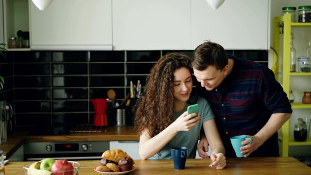 young-attractive-curly-happy-caucasian-woman-is-sitting-in-kitchen-texting-someone-via-smartphone,-her-husband-comes-and-she-shows-something-funny-in-phone