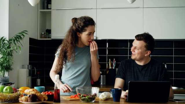Cheerful-couple-at-table-woman-dancing-and-cooking-while-gives-hasband-to-try-red-pepper,-man-sitting-working-on-laptop,-he-shows-something-funny-on-screen