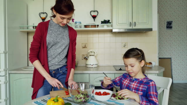 Happy-young-mother-and-cheerful-daughter-cook-salad-together-in-the-kitchen-at-home-cutting-vegetables-and-talking.-Family,-cooking,-and-people-concept