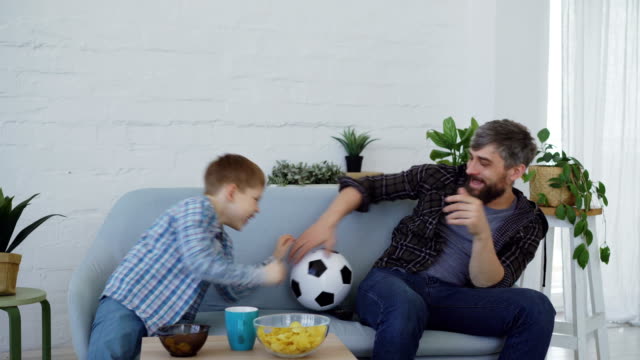 Entzückende-Geißlein-ist-mit-Ball-mit-seinem-aktiven-fürsorglichen-Vater-spielen,-werfen-und-fangen-Fußball-auf-Couch-zu-Hause.-Familie-Kleben-und-lustigen-Sport-Konzept.