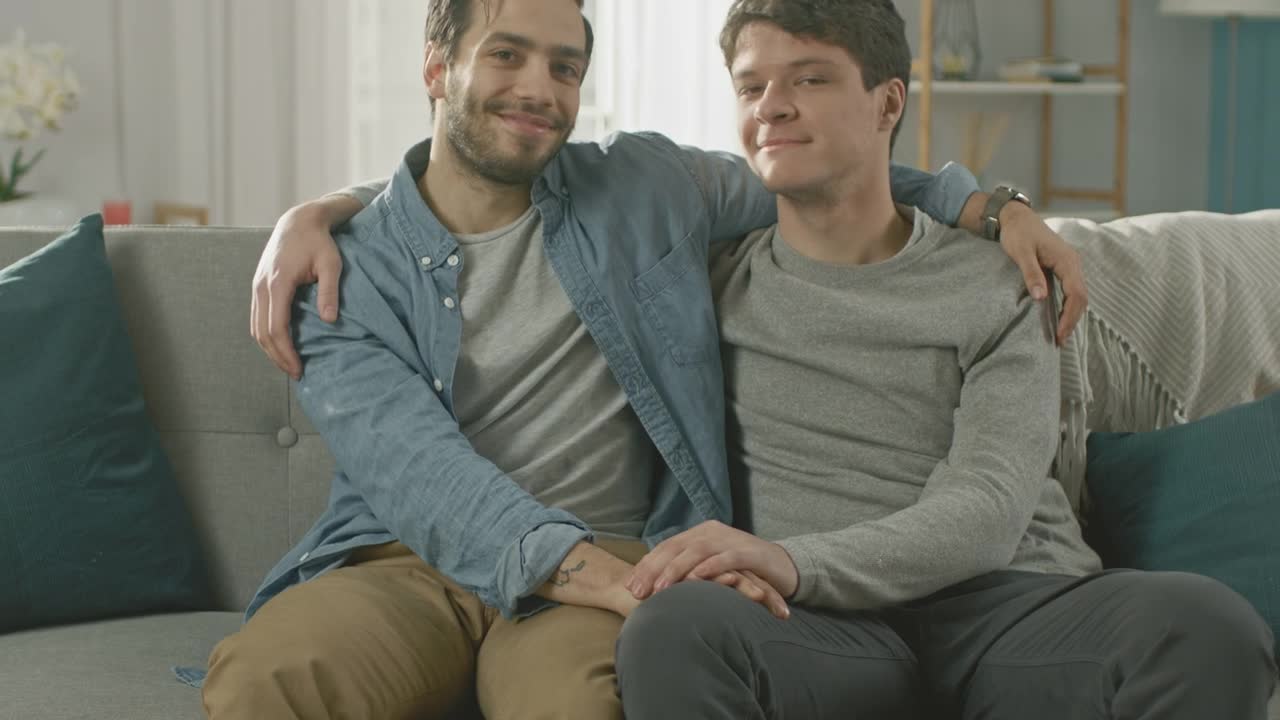 Close Up Shot Of A Cute Male Gay Couple Sitting Together On A Sofa At Home.  They Are Hugging And Holding Hands. They Are Happy And Smiling. They Are  Casually Dressed And