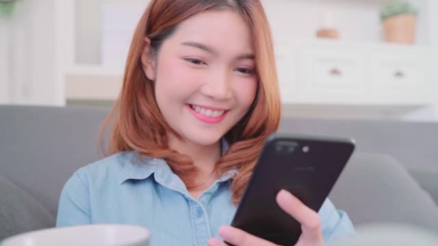 Portrait-of-beautiful-attractive-smiling-Asian-woman-using-smartphone-holding-a-warm-cup-of-coffee-or-tea-while-lying-on-the-sofa-when-relax-in-living-room-at-home.-Lifestyle-women-at-home-concept.