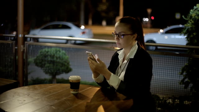 hermosa-mujer-caucásica-joven-sentada-después-del-trabajo-por-la-noche-en-un-café-en-la-calle-bebiendo-café-y-charlando-en-las-redes-sociales-usando-un-smartphone.