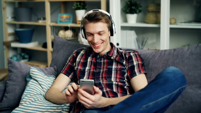 Glücklicher-junger-Mann-in-Kopfhörern-ist-Musik-zu-hören-und-mit-Smartphone-zu-Hause