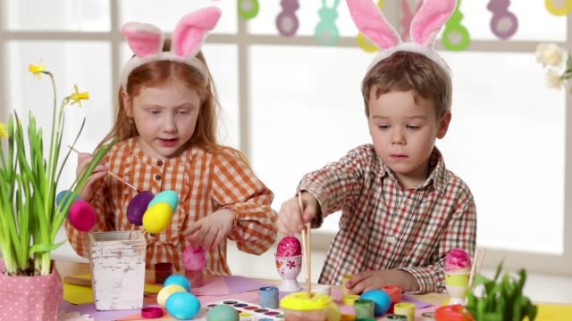 Niños-felices-usando-orejas-de-conejo-pintando-huevos-el-día-de-Pascua.-Niñas-pequeñas-preparándose-para-la-Pascua.