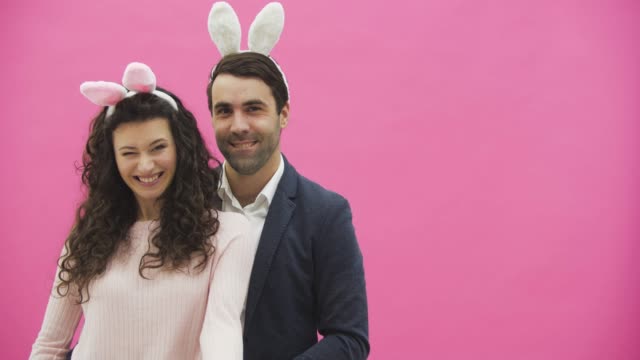 Young-couple-standing-on-a-pink-background.-During-this,-they-look-at-the-camera-and-show-a-gesture-of-call.-Happy-family-is-preparing-for-Easter,-with-the-ears-of-a-pink-rabbit-on-its-head.