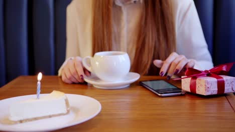 Unrecognizable-red-haired-woman-sitting-in-cafe-and-browsing-cell-phone-on-her-birthday.-Cup-of-tea,-gift-box-and-piece-of-cake-with-burning-candle-on-her-table