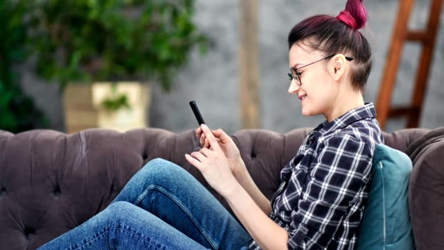 Smiling-hipster-young-woman-chatting-using-smartphone-sitting-on-couch-at-home-medium-shot