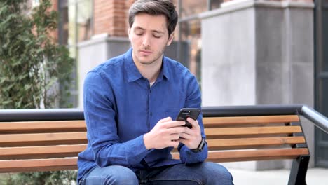 Junger-Mann-tippen-Nachricht-auf-Smartphone,-sitzen-draußen-auf-Bank