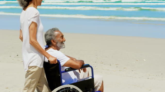 Vista-lateral-de-la-mujer-afroamericana-activa-Senior-con-el-hombre-mayor-discapacitado-en-la-playa-en-sol-4k