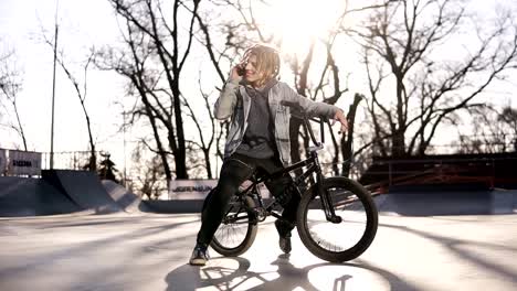 Retrato-de-feliz-adolescente-guapo-con-rastas,-utilizando-el-teléfono-móvil-mientras-se-monta-una-bicicleta-BMX,-sonriendo,-amigos-de-mensajería-a-través-de-las-redes-sociales.-Personas,-estilo-de-vida-y-concepto-de-comunicación.-El-metraje-de-ángulo