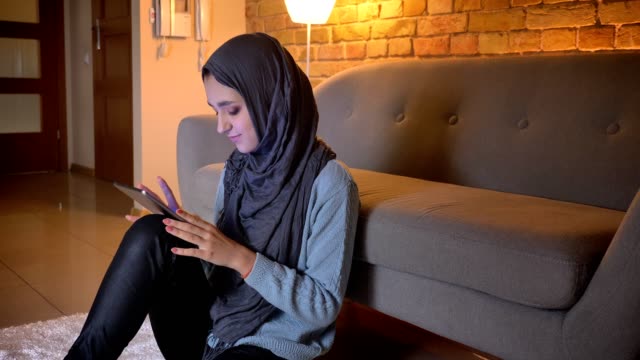 Closeup-shoot-of-young-attractive-muslim-female-smiling-and-browsing-on-the-phone-while-sitting-on-the-floor-indoors-at-cozy-home