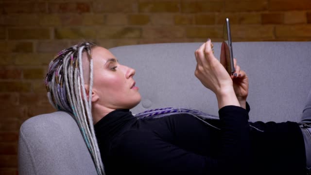 Closeup-shoot-of-young-attractive-caucasian-hipster-female-using-the-tablet-turning-to-camera-and-happily-smiling-lying-on-the-couch-indoors-in-a-cozy-apartment