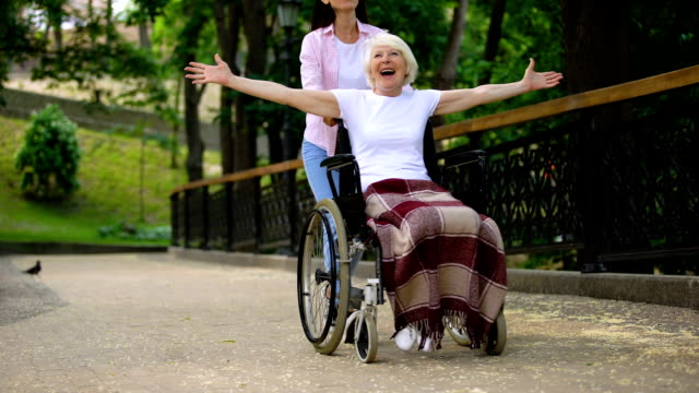Glückliche-Freiwillige-schieben-Rollstuhl-mit-positiven-alten-Dame,-fröhliche-Patientin