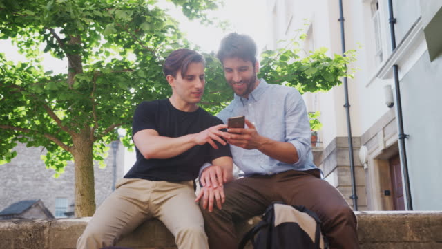 Männliche-Homosexuell-Paar-sitzen-im-Freien-an-der-Wand-des-Gebäudes-Blick-auf-Handy