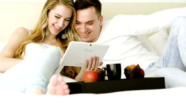 Happy-couple-watching-a-funny-movie-on-tablet-computer-together-in-bedroom.-4K