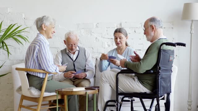 Gruppe-von-vier-älteren-Menschen,-darunter-behinderte-Männer-im-Rollstuhl,-die-sprechen,-während-sie-gemeinsam-im-Pflegeheim-Karten-spielen