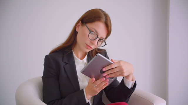 Nahaufnahme-Porträt-der-jungen-kaukasischen-Geschäftsfrau-in-Brille-tippen-auf-dem-Tablet-aussehenta-Kamera-lächelnd-glücklich-sitzend-im-Sessel