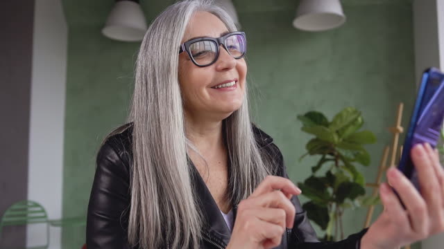 Close-up-of-beautiful-gray-hair-smiling-woman-in-black-jacket-waving-her-hand-to-the-interlocutor-while-having-video-call-at-home