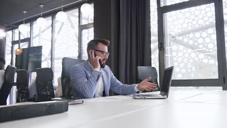 Serious-high-skilled-businessman-discussing-computer-project-on-phone-with-interlocutor-sitting-at-the-office-workplace