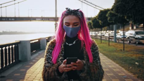 Joven-mujer-hipster-con-ropa-informal-y-máscara-protectora-está-escribiendo-en-el-teléfono-celular,-caminando-por-terraplén