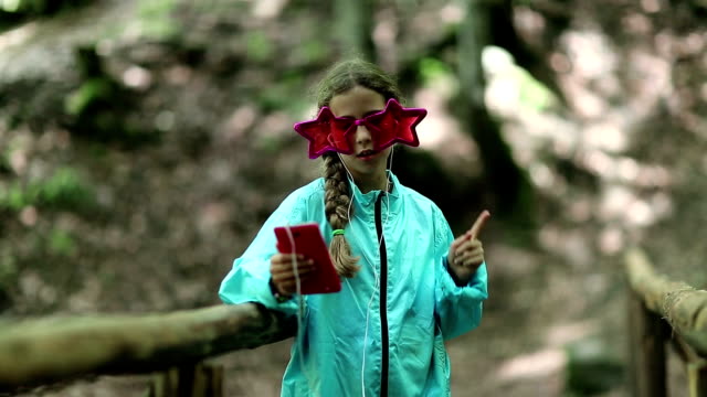 Girl-with-red-smartphone-stands-on-wooden-bridge,-listens-to-music-and-dance